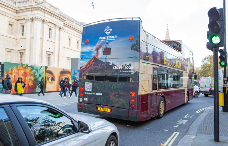 the focus of the picture is a double-decker bus driving through the city of London with a big advertisement for Guatemala on its rear side.