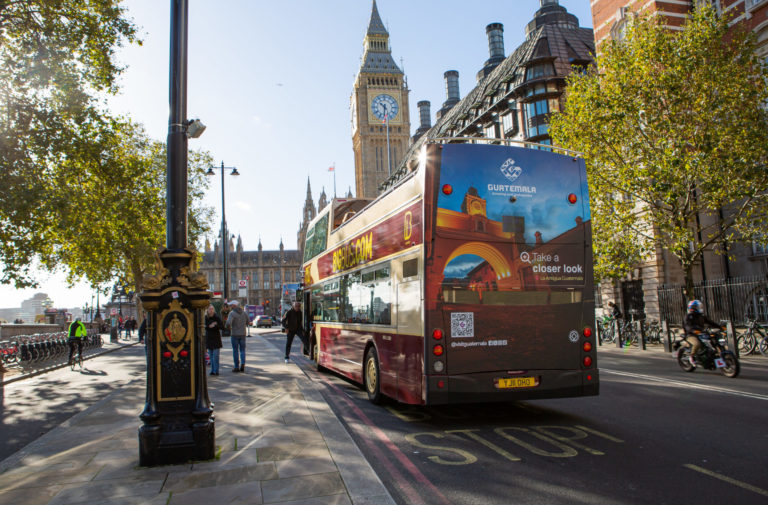 A large bus drives through London on a sunny day. On the back is a huge advertisement for the destination "Guatemala".