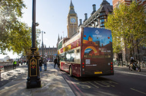 A large bus drives through London. The sun is shining and on the rear side, there is a big advertising for a destination called "Guatemala"