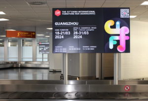 Over luggage belt at the Cologne airport is a digital video board showing an ad for Chinas International Furniture Faire (CIFF). Around the out-of-home advertising there are other luggage belts, escalators, and signs visible.