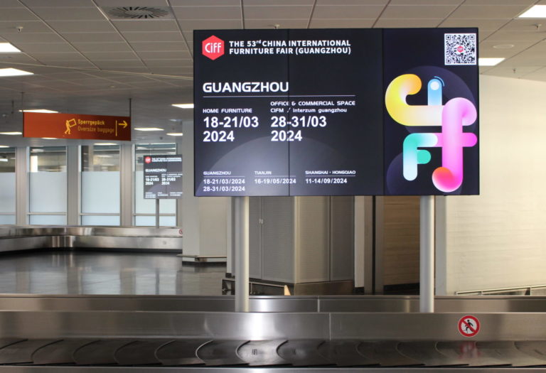 Over luggage belt at the Cologne airport is a digital video board showing an ad for Chinas International Furniture Faire (CIFF). Around the out-of-home advertising there are other luggage belts, escalators, and signs visible.