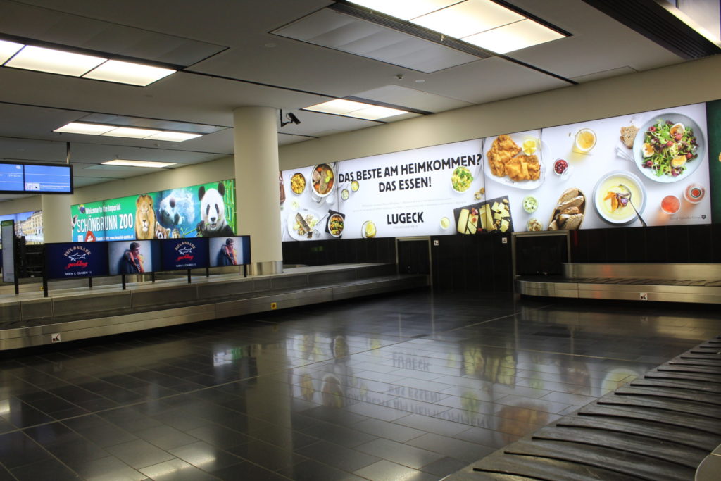 The picture shows Vienna Airport with a baggage carousel on which several screens are placed. In the background is a large screen showing advertising for Lugeck.