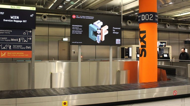 Over the luggage belt on video boards at the Cologne airport the CIFF celebrates their 55th anniversary. Around the video board is the wating area for the baggage claim. People are standing at the belt.