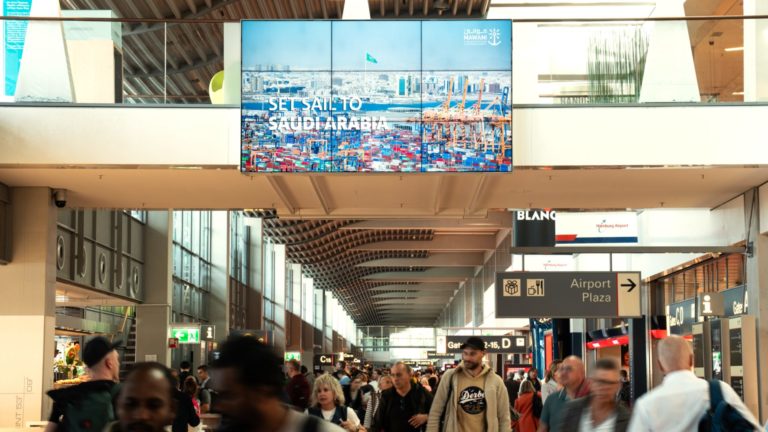 Over a passageway at the Hamburg airport hangs a digital video wall showing the advertisement of Mawani. You can see the slogan “Set to Sail to Saudi Arabia” in combination with a picture of the their port. Below the airport advertising are people passing through. A long side the way are many stores.