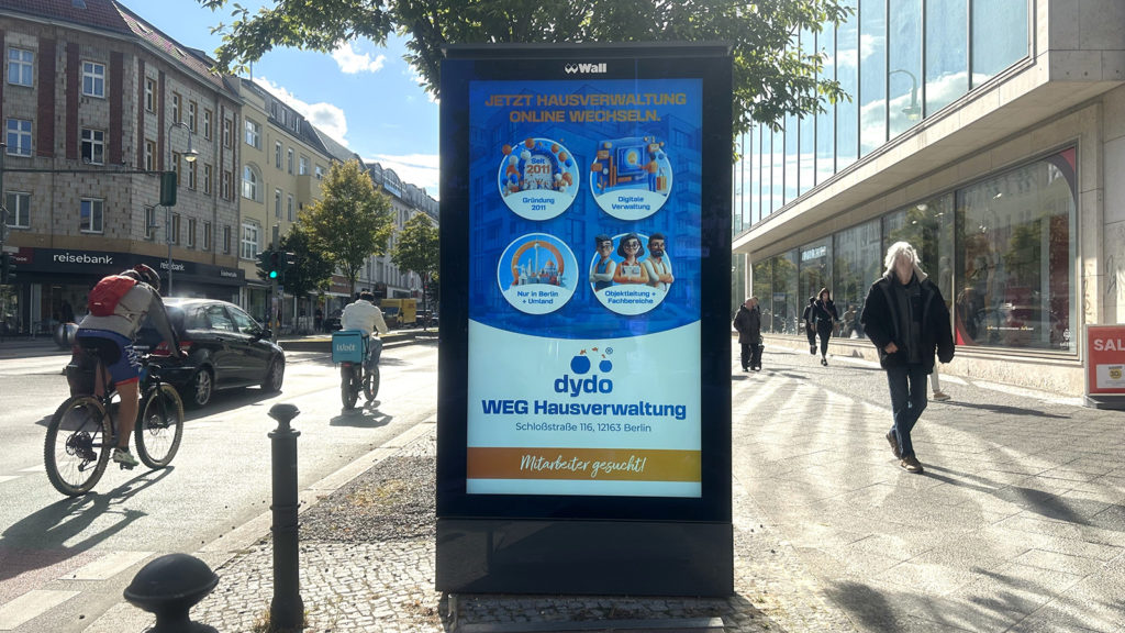 At the center is a digital city light poster featuring an advertisement for Dydo. On the left side of the image, a road with passing cars and a cyclist is visible. On the right, a pedestrian is walking by.