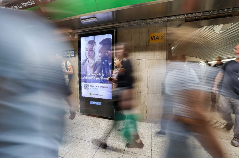 Digital city light poster in the metro in Barcelona, with passengers walking by around it.