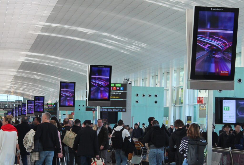 The busy arrivals area at Barcelona Airport. You can see a large number of city light posters connected in series, mounted above people's heads. The motif on the digital display, advertising the company Barco, is futuristic and in shades of purple and blue.