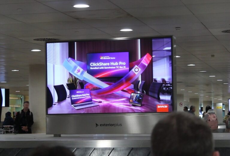 The baggage area at Barcelona Airport. You can see people waiting for their luggage. A large digital advertising screen in landscape format is located directly above one of the baggage carousels. The advertisement is designed in bold, eye-catching colors and shows Barco's new video conferencing system.