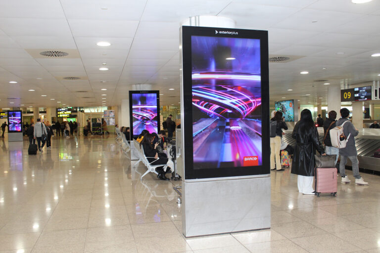 The baggage claim at Barcelona Airport, where many people are waiting for their luggage. Among the waiting passengers, three vertically arranged digital screens are visible. The digital display is running a spot for the company Barco.