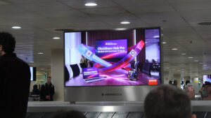 The baggage claim at Barcelona Airport. Passengers are waiting for their luggage. Directly above one of the carousels is a large, wide-format digital advertising screen. The advertisement is designed in bold, striking colors and showcases Barco’s new video conferencing system.