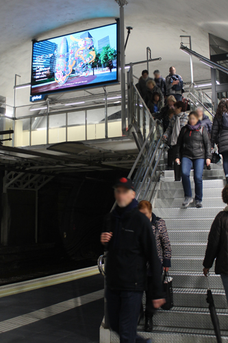 A staircase serving as a footbridge directly above a railway tunnel. Many people are walking up and down it. In the middle, a large digital billboard hangs from the ceiling, displaying a brightly colored, futuristic design for the ISE trade show.