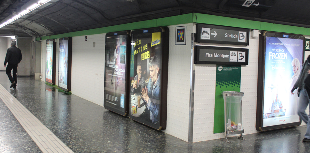 A corridor in a subway station. Five vertical advertising panels hang on the walls in display cases, each featuring a different colorful, backlit poster. Between the posters, there is a sign pointing to the Fira Montjuïc exhibition center.