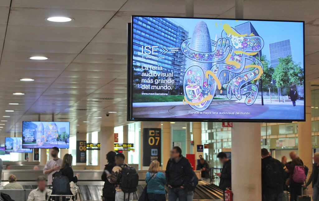 The baggage claim at Barcelona Airport with waiting passengers. Above the carousels there are three large, wide-format digital advertising screens. On the front screen, between the buildings of Barcelona, a fantastical, futuristic figure is visible along with a slogan promoting the ISE trade show in Barcelona.