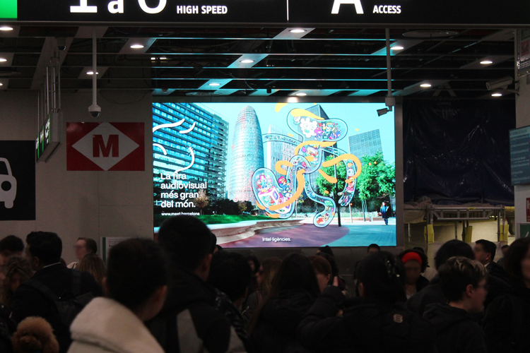 Hall A at Barcelona’s main train station with a crowd of people. A large digital billboard in landscape format hangs on the wall. On the screen, between Barcelona’s buildings, you can see an imaginative, futuristic creature and a slogan promoting the ISE trade show.