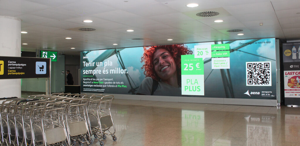 A very large wide-format digital advertising screen mounted on the wall at Barcelona Airport. Luggage carts are in the foreground.