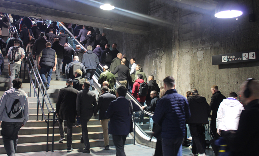A station staircase with many people walking up the stairs. On the right-hand wall, there is a sign pointing the way to the exit and to the Fira Gran Via exhibition grounds.