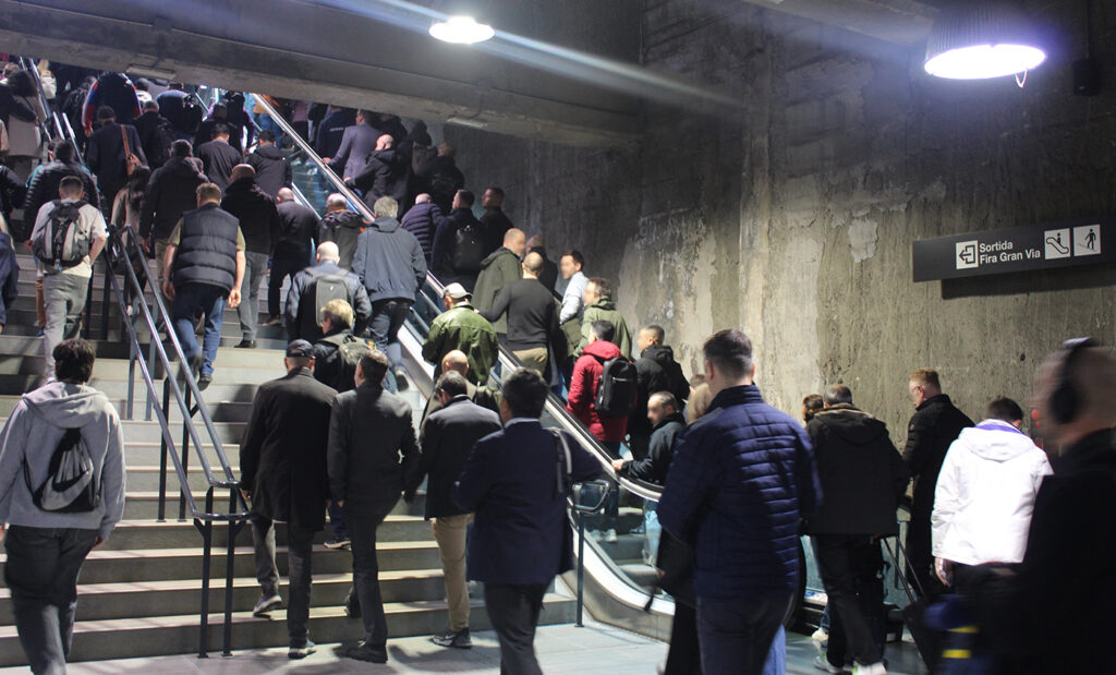 Interior view of a train station with a staircase and an escalator side by side, many people walking upward. A signpost reading “Fira Gran Via” hangs on the wall to the right.