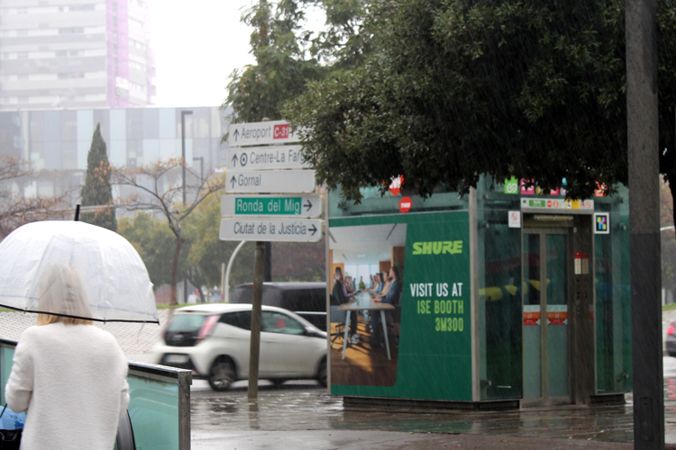 A station plaza next to a street. To the left is the escalator leading to the basement; to the right is an elevator. The elevator is completely covered in outdoor advertising. The company Shure is promoting its trade show booth, using a green design and an image of a conference.