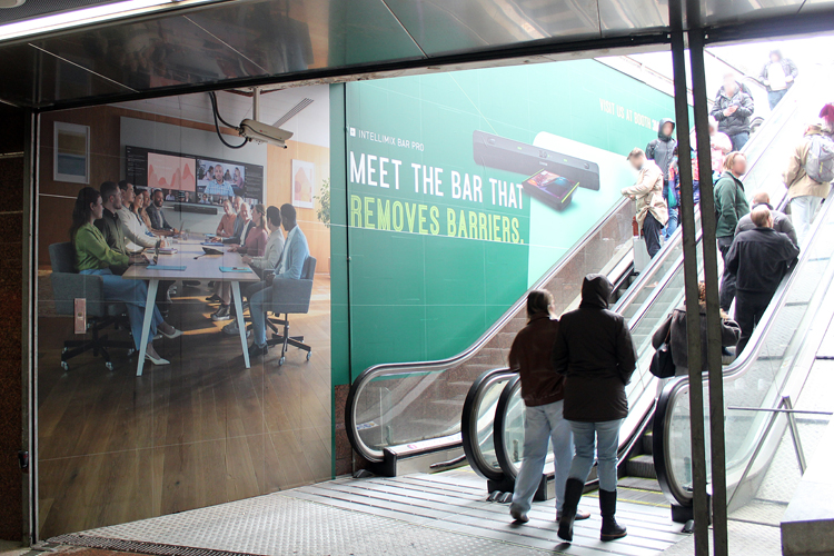 An escalator in the train station leading outside. Many people are riding it up. A large advertisement is posted on the wall right next to the escalator. The company Shure is promoting its new video bar for online conferences. The advertisement is green, featuring text and images of an office and the video bar.