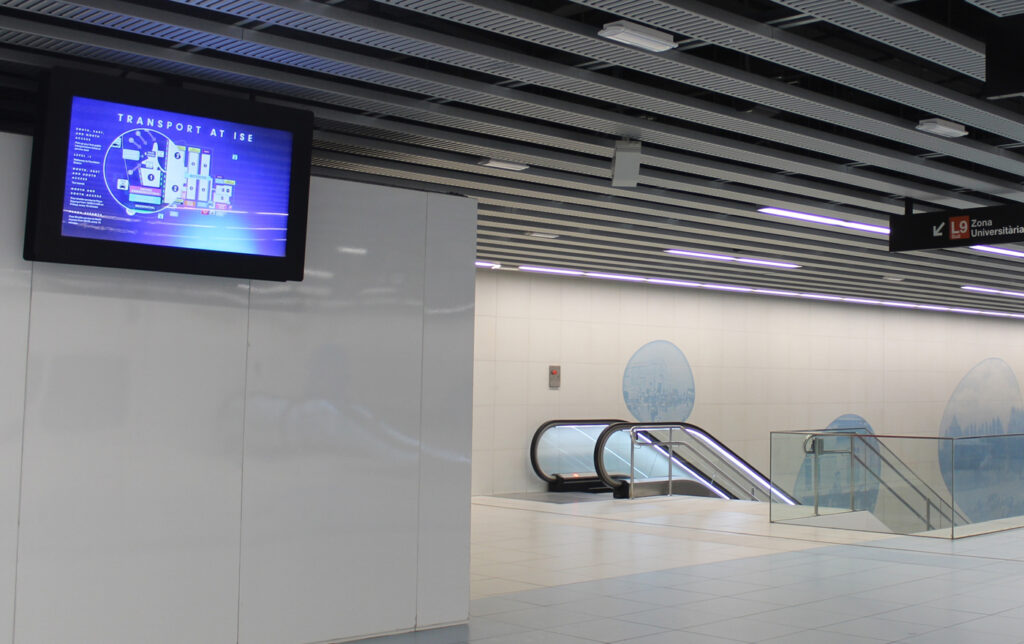 A light blue station concourse with an escalator leading down to the basement. On the left side, a landscape-format digital advertising screen hangs from the ceiling. It displays directions to the ISE trade show in a blue design.