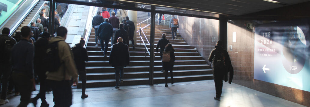 A set of stairs at the train station leading outside. Many people are walking up the stairs. On the right-hand wall, there is a large blue poster pointing the way to the ISE 2026 trade show.