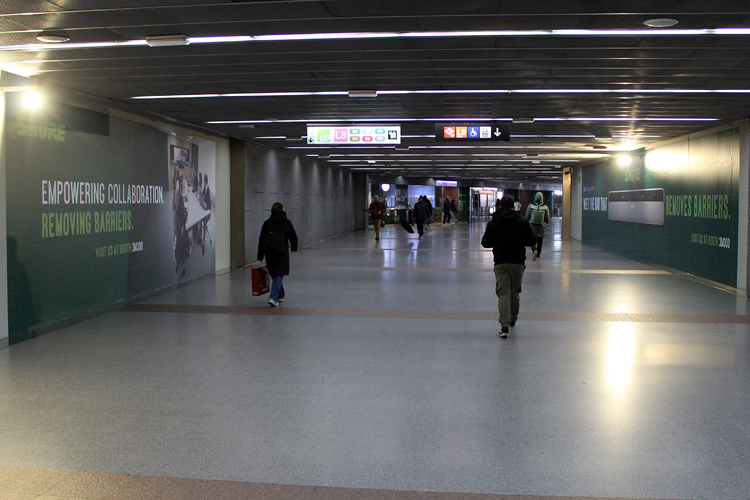 A walkway in a subway station with people. On the wall of the walkway are two large, floor-to-ceiling, and very wide advertising spaces featuring a green design by the company Shure.