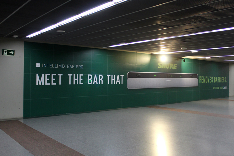 A large, floor-to-ceiling, and very wide advertising space on the wall of the subway walkway. The company Shure is promoting its newly developed video bar for meetings. The company uses a green design featuring text and an image of the video bar.