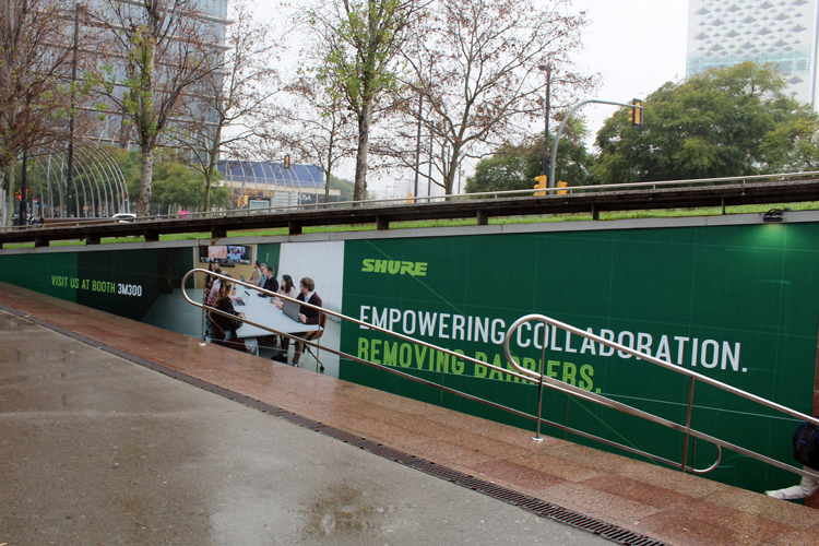 A staircase leading from the train station to the street level. The side wall of the staircase is completely covered with an advertisement. With a green background, Shure uses text to promote its trade show booth and its new product, a video bar for online conferences.