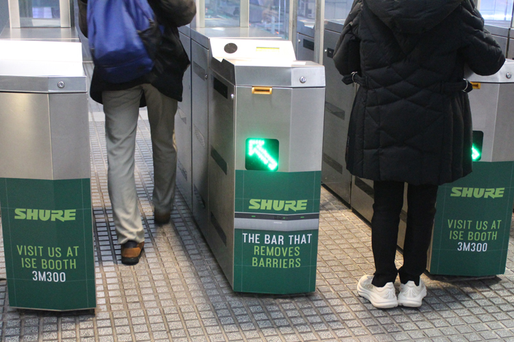 Two People standing in front of three ticket-checking machines at the subway station. The lower half of the machines is covered with advertisements. The company Shure is promoting its trade show booth with green-colored lettering and images of its new video bar for online conferences.