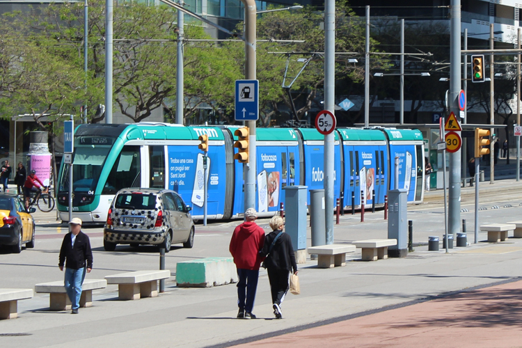 In the foreground, people are walking and cycling along a pedestrian and bike path. In the background is a street with cars and a tram. The tram is completely covered in outdoor advertising. The design is blue and turquoise.