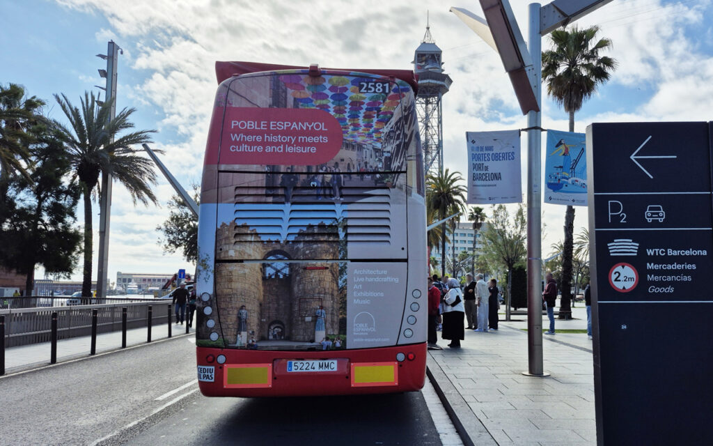 A bus on the street right next to the Barcelona coastline. You can see the back of the bus, which is completely covered with an advertising wrap. The advertisement features Poble Espanyol, a Barcelona landmark.