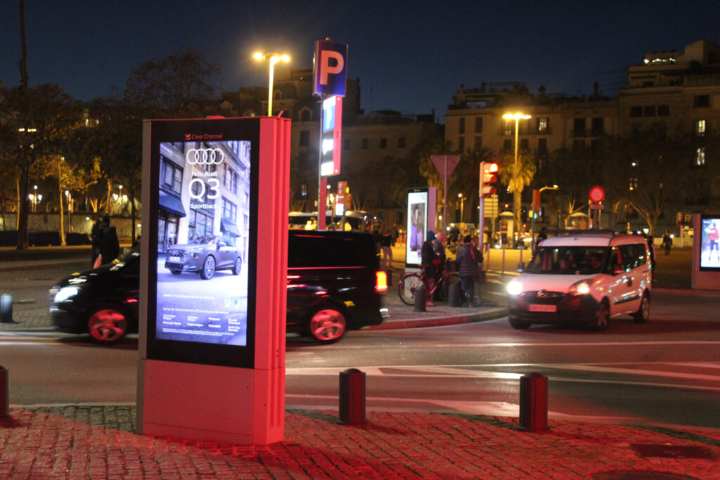 A nighttime photo showing a digital City Light Poster at a busy intersection, with pedestrians and cyclists on the sidewalk. In the background are two more digital City Light Posters.
