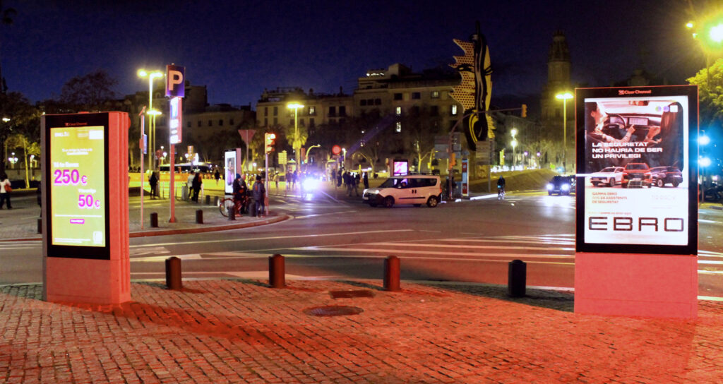 A nighttime shot of two digital City Light Posters at a bustling intersection filled with cars, pedestrians, and scooter riders. The ads on the displays show two different images: one for cars and one for a bank.