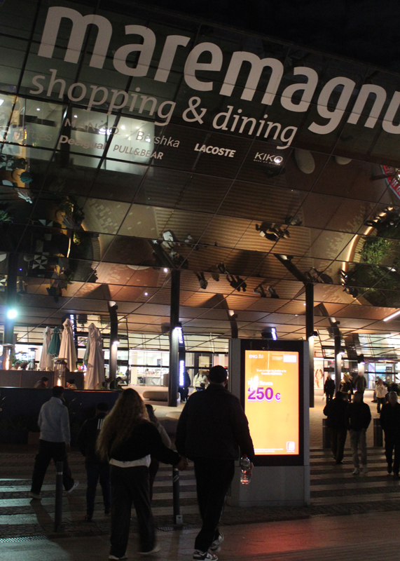 A nighttime shot of the entrance to a large shopping center, with many people streaming up the stairs toward the doors. In front of the stairs there is a digital City Light Poster advertising a bank.