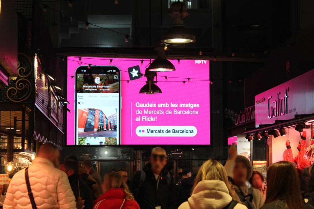 A market hall with stalls on both sides. A crowd of people is walking down an aisle between the stalls. At the end of the aisle, a large digital billboard hangs on the wall. The advertisement is a striking shade of pink.
