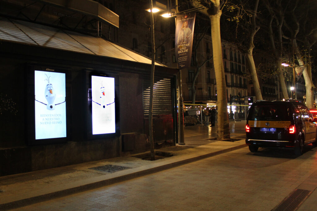 A street with cars, night shot. Two bright, digital City Light Posters stand out; they display an advertisement for Disney's Frozen, featuring mostly white and light blue colors.