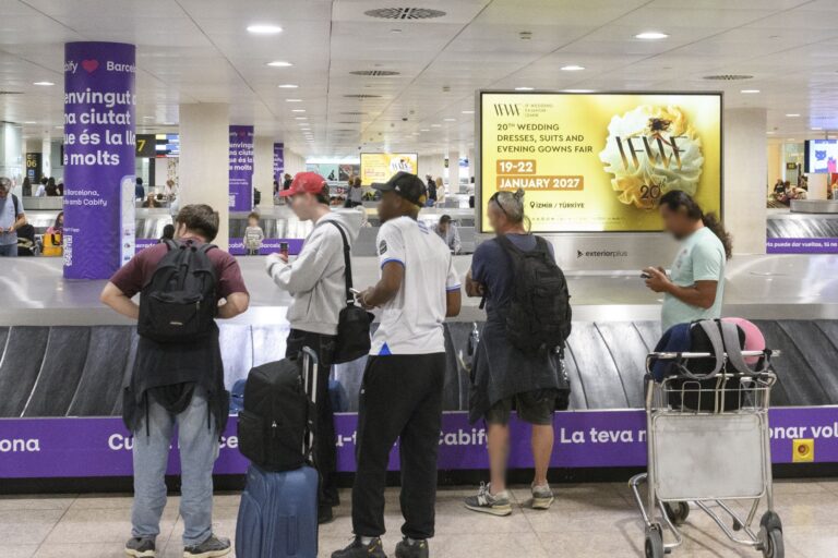In the baggage claim area of the airport, digital advertising displays in landscape format are installed in a row. Numerous passengers are standing nearby, waiting for their luggage. An advertising campaign is running on the screens for a bridal fashion trade fair taking place in Izmir in January 2027.