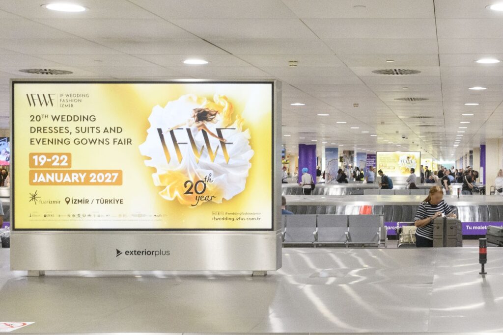 Several landscape-format digital advertising screens installed in a row in the airport baggage claim area. Many people can be seen waiting for their luggage. The running advertisement is designed in a striking yellow and promotes a bridal fashion trade show in Izmir in January 2027.