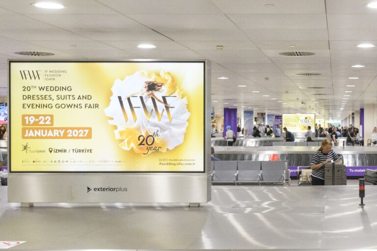 Several landscape-format digital advertising screens installed in a row in the airport baggage claim area. Many people can be seen waiting for their luggage. The running advertisement is designed in a striking yellow and promotes a bridal fashion trade show in Izmir in January 2027.