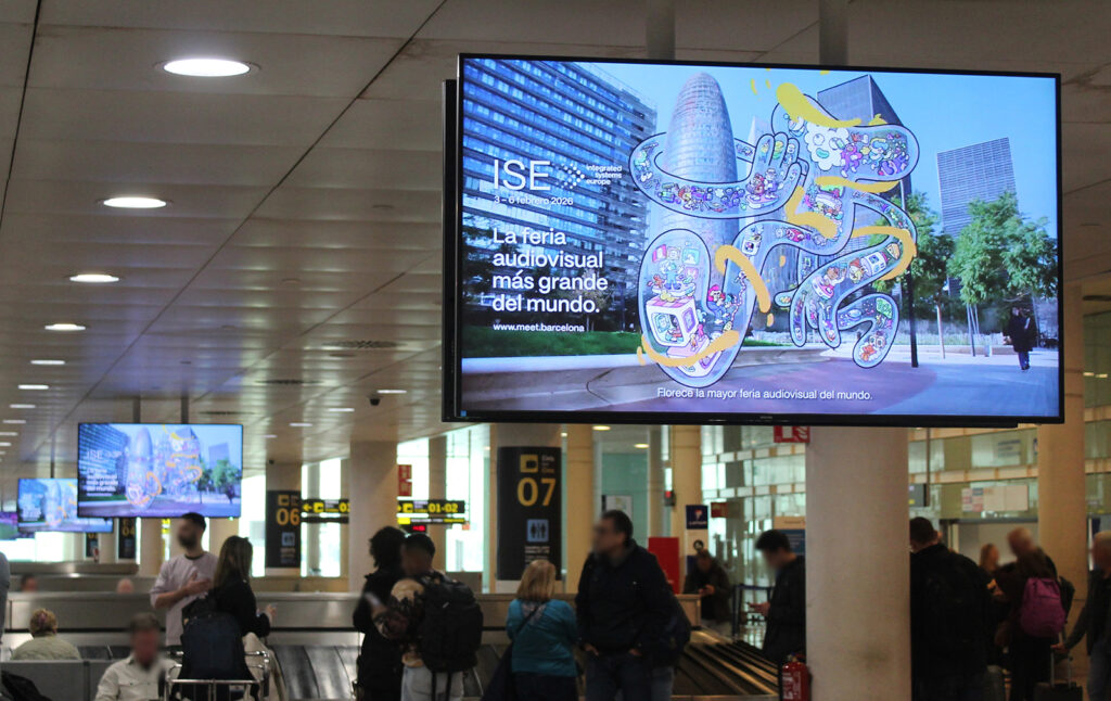 Several large-format digital advertising screens in the baggage claim area at the airport. The screens display advertisements for the ISE trade show. People are waiting for their luggage.