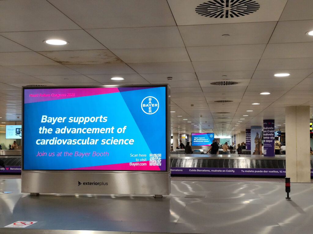 Large digital advertising displays in the airport’s baggage claim area show advertising for the trade fair appearance from the company Bayer. Passengers wait to collect their luggage.