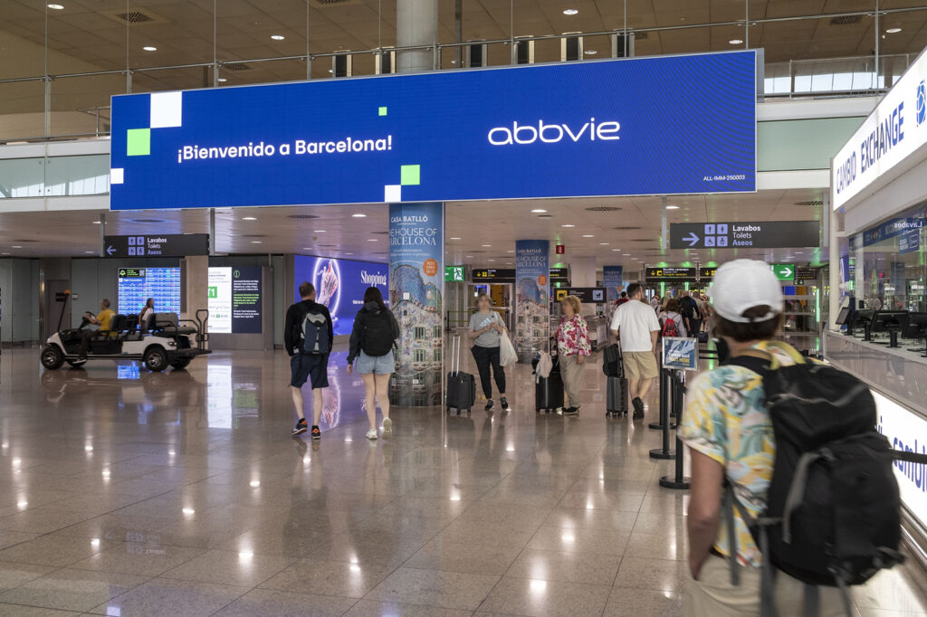 A large-format digital video wall at the airport. The display shows a blue advertisement from AbbVie.