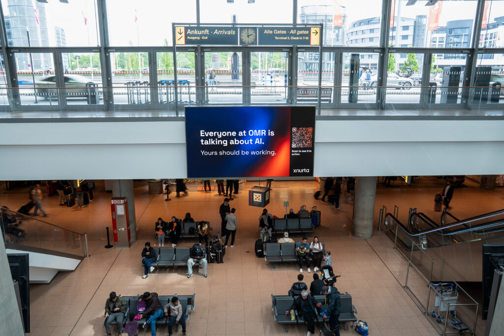 Digital screen mounted at the ceiling in an airport underground waiting area. The screen shows a large orange and blue ad from the brand Xnurta for the OMR trade show. People sit on benches and walk through the space below.