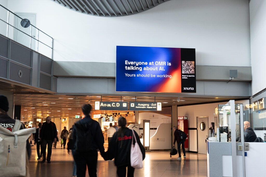 A digital advertising screen in the airport terminal, with passengers walking beneath it in the passageway. The screen displays an advertisement for Xnurta featuring the text “Everyone at OMR is talking about AI. Yours should be working,” along with a QR code.