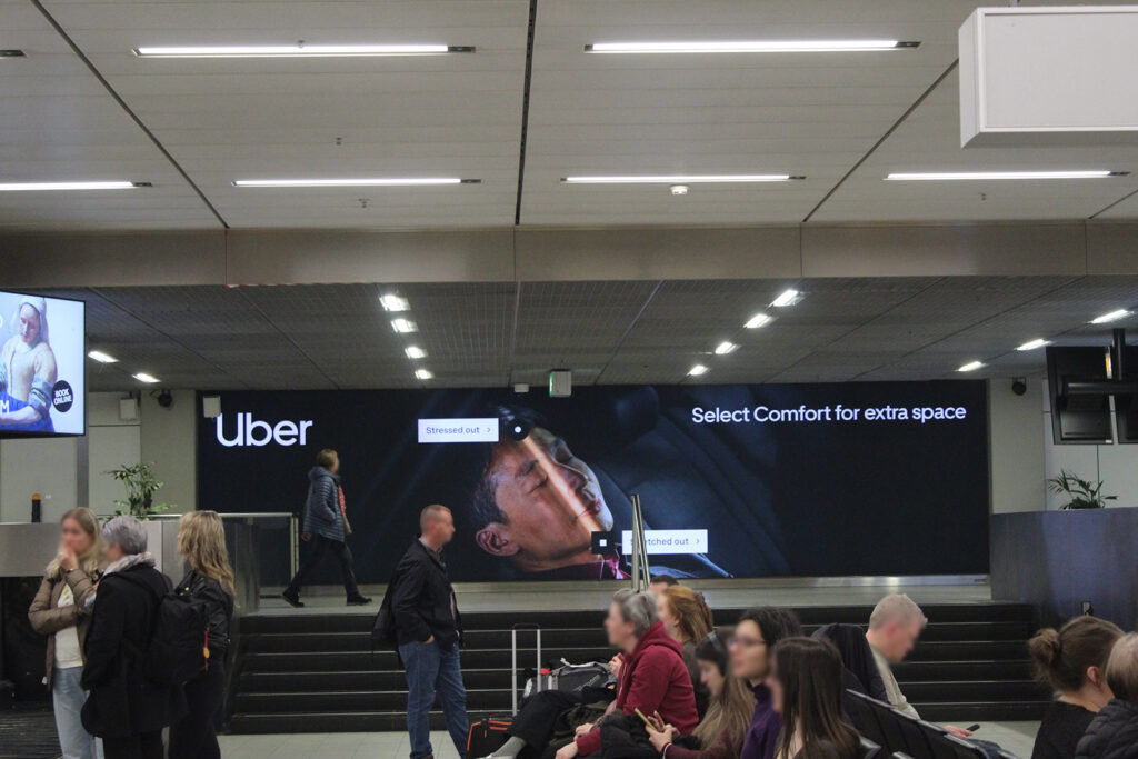 A large landscape format backlit lightbox in a baggage hall at Amsterdam Airport. The advertisement is for Uber. People are sitting on benches.