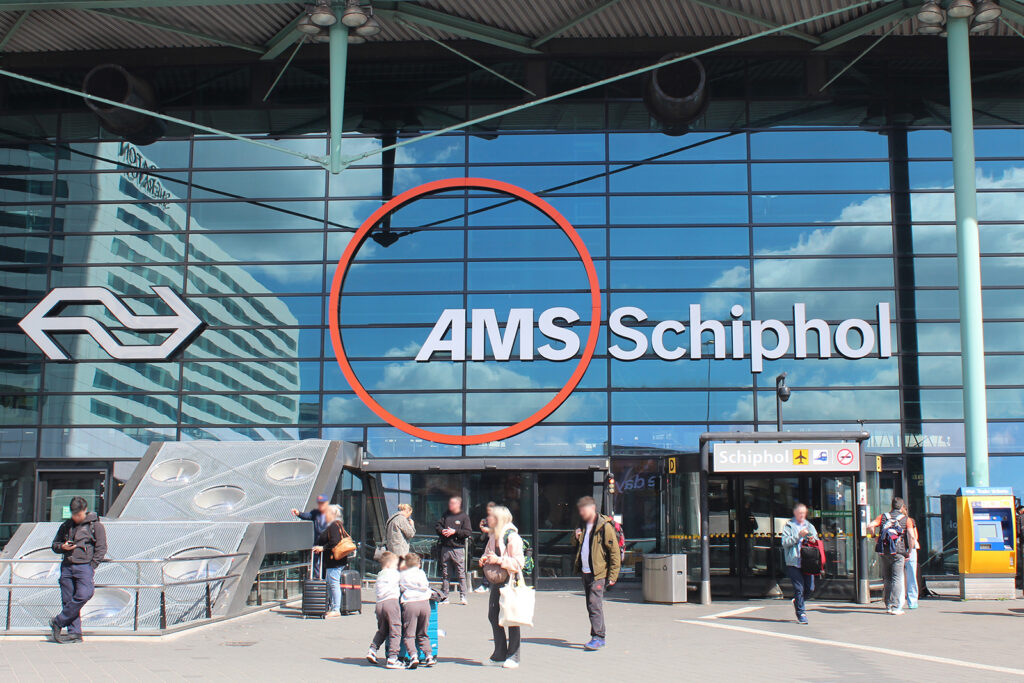 The entrance to Amsterdam Schiphol Airport. People are gathered outside the terminal, the building features a glass facade, and a large Schiphol Airport logo is displayed on the windows.