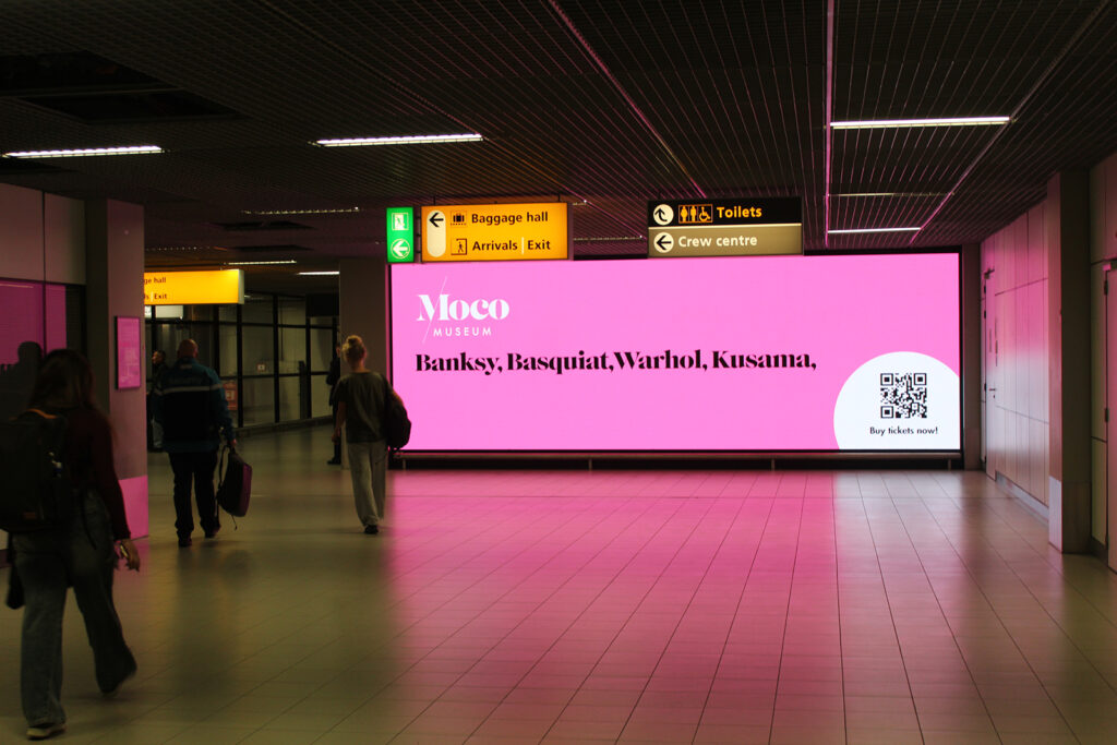 A large digital advertising screen in the airport corridor. People are walking past the digital screen. The screen displays pink advertising for the Moco Museum in Amsterdam.