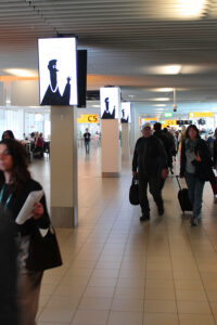 Several portrait format digital City Light Posters on multiple columns at Amsterdam Airport. Two screens are mounted on each column with their backs facing each other. People are walking past the illuminated advertising displays.