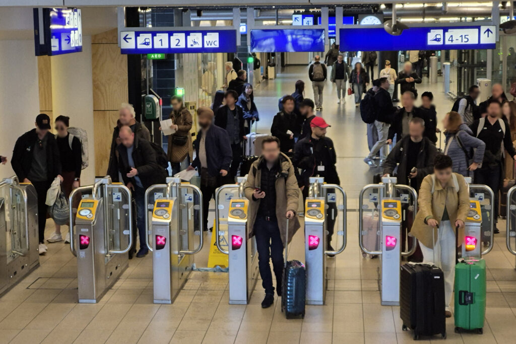 The main concourse of Amsterdam Central Station. Numerous people with and without luggage are moving through the hall. Some are scanning their tickets, while others are walking or waiting.