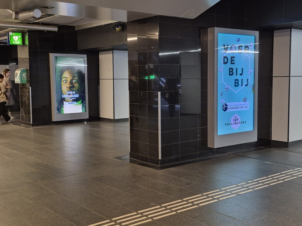 Digital City Light Posters at Amsterdam Central Station. They are mounted on the walls and illuminated. People walk past them.